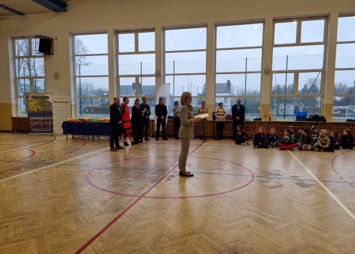 Zdjęcie 1. Przywitanie uczestników spotkania przez Panią Justynę Jakubik z ZSP w Otrębusach. Od lewej na dalszym planie znajdują się: Pan Sebastian Kończak Dyrektor Zespołu Szkolno – Przedszkolnego w Otrębusach, Pan Marcin Błaszczyk – Ratownik Medyczny z SPZOZ „MEDITRANS” w Warszawie, Pani Magdalena Kalniuk – koordynatorka Kampanii p.n. „JWJB” z  Urzędu Marszałkowskiego Województwa Mazowieckiego  w Warszawie, Pan Sławomir Bakalarski – Egzaminator z Wojewódzkiego Ośrodka Ruchu Drogowego w Warszawie, Starszy Sierżant Piotr Ostrowski - Policjant z Komendy Powiatowej Policji w Pruszkowie. (zdjęcie źródło: WORD w Warszawie). 