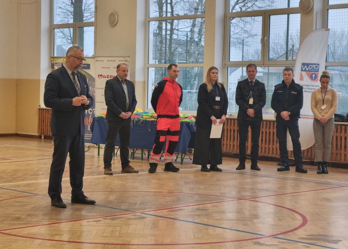 Zdjęcie 3. Na zdjęciu (pierwszy od lewej) Pan Jerzy Wysocki – Drugi Zastępca Burmistrza Gminy Brwinów podczas przemówienia do uczestników spotkania. Na dalszym planie znajdują się (od lewej): Pan Sebastian Kończak Dyrektor Zespołu Szkolno – Przedszkolnego w Otrębusach, Pan Marcin Błaszczyk – Ratownik Medyczny z SPZOZ „MEDITRANS” w Warszawie, Pani Magdalena Kalniuk – koordynatorka Kampanii p.n. „JWJB” z  Urzędu Marszałkowskiego w Warszawie, Pan Sławomir Bakalarski – Egzaminator z Wojewódzkiego Ośrodka Ruchu Drogowego w Warszawie, Starszy Sierżant Piotr Ostrowski - Policjant z Komendy Powiatowej Policji w Pruszkowie, Pani Kamila Michalak-Jobda z Delegatury Urzędu Marszałkowskiego Województwa Mazowieckiego w Piasecznie. (zdjęcie źródło: WORD w Warszawie).